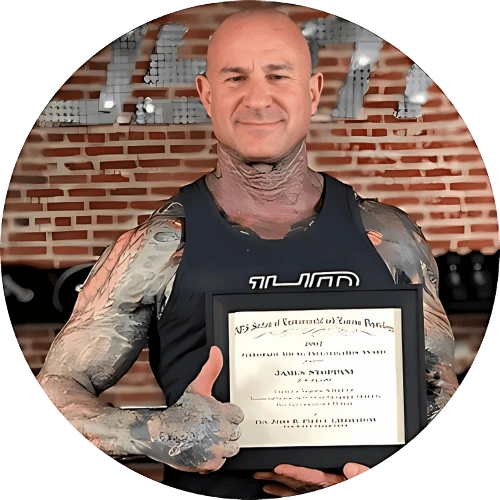 A muscular, tattooed man in a sleeveless shirt poses with a framed certificate in front of a brick wall background. He smiles and gives a thumbs-up with one hand while holding the certificate from Dr. Stoppani with the other.