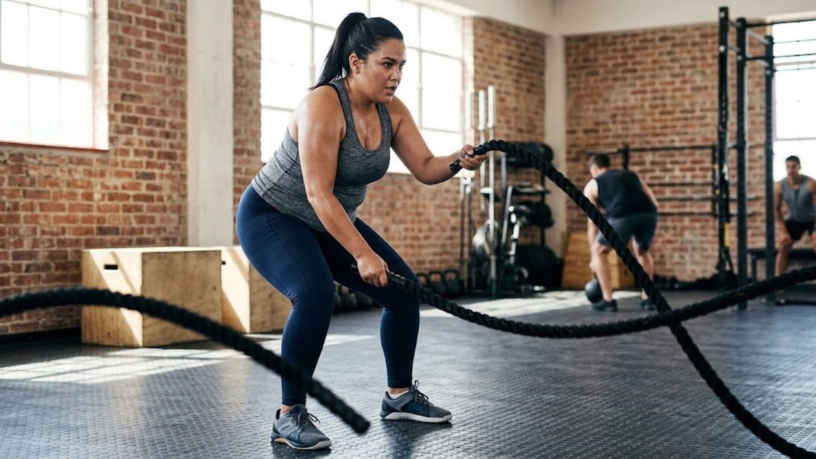 Woman in a gym using battle ropes, wearing a gray tank top and blue leggings with sunlight streaming through brick walls in the background.